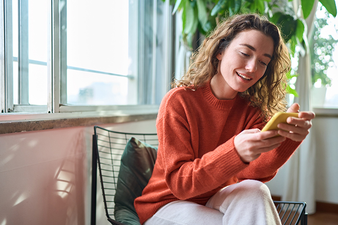 Smiling young woman sitting on chair holding mobile phone using cell at home., Smiling young woman sitting on chair holding mobile phone using cellphone device, looking at smartphone, checking modern apps, texting messages, browsing internet doing shopping relaxing at home.