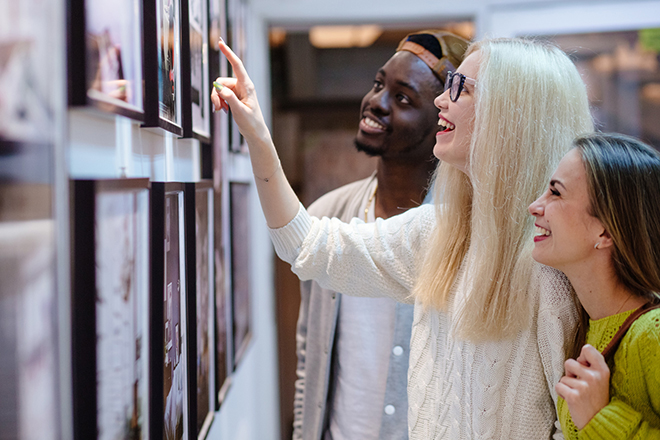 Three young people hipster students with different skins looking happy together, talkng, looking at picture on wall, spending free time together in art photo gallery. Multi ethnic, friendship concept.